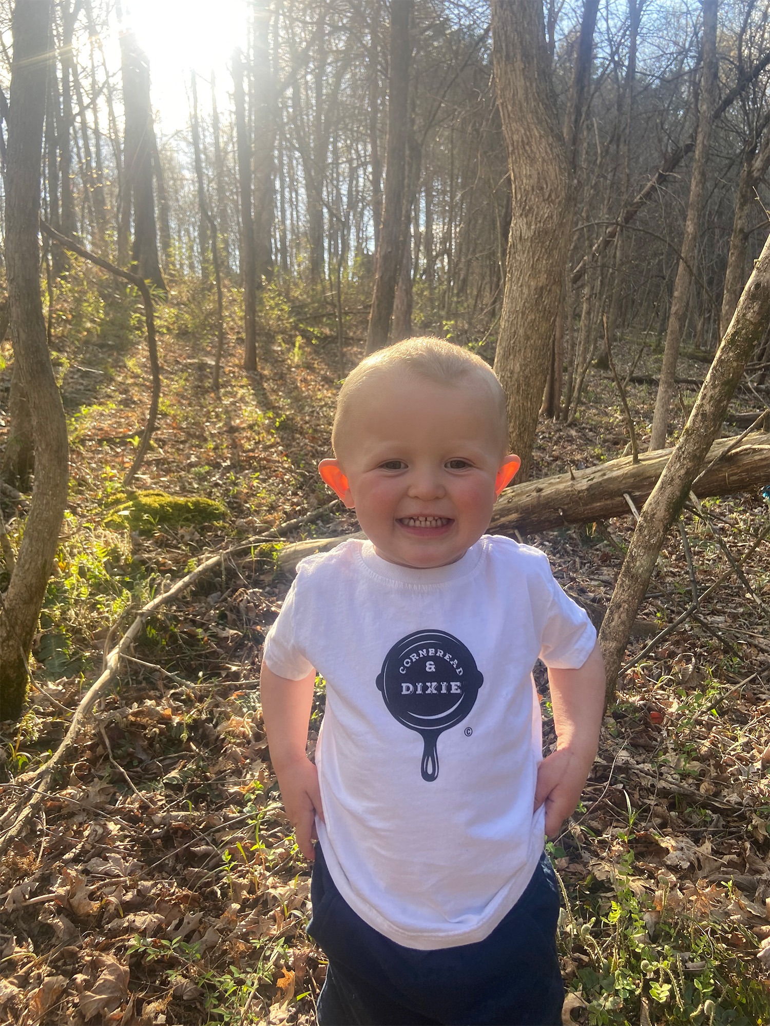 Smiling toddler outdoors in a forest, wearing a white shirt with "Cornbread & Dixie" logo. Sunlight filters through the trees, illuminating the scene.