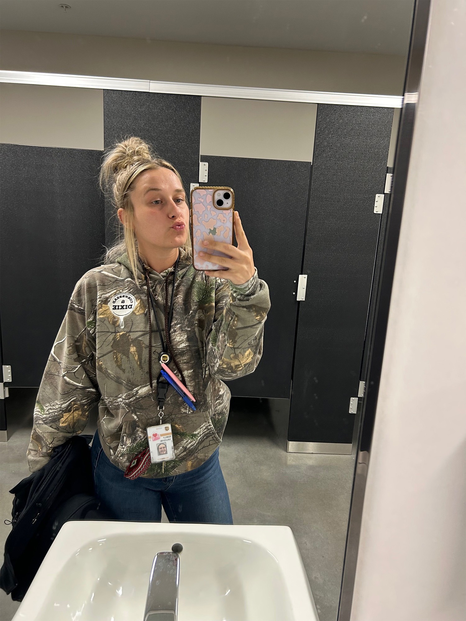 Young woman in a camouflage hoodie poses for a selfie in a bathroom mirror, holding a phone and displaying a work ID.