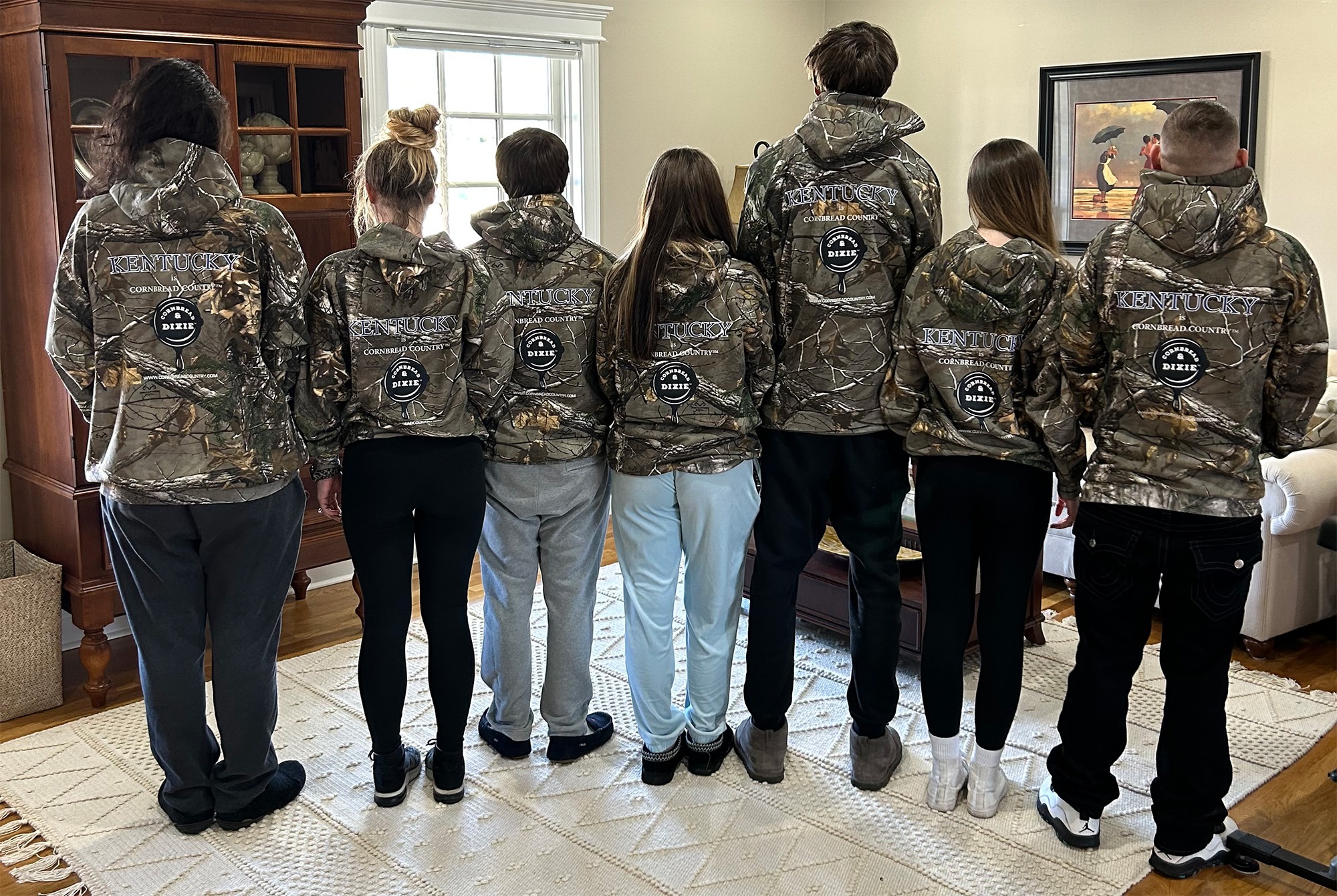 A group of six individuals stands side by side, wearing matching camouflage jackets with "Kentucky Cornbread Country" branding, facing away from the camera.