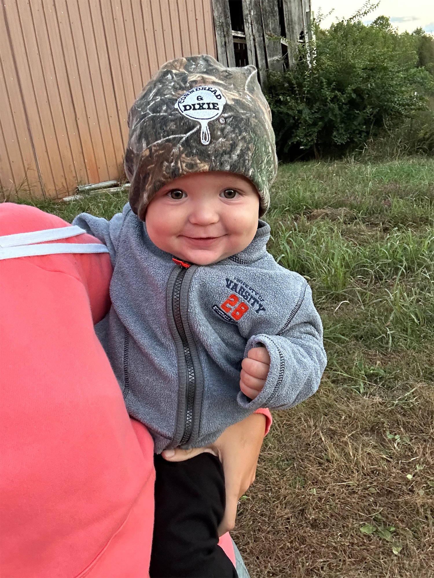 Smiling toddler in a camouflage hat, being held by an adult in a pink sweatshirt, set against a rustic barn backdrop.