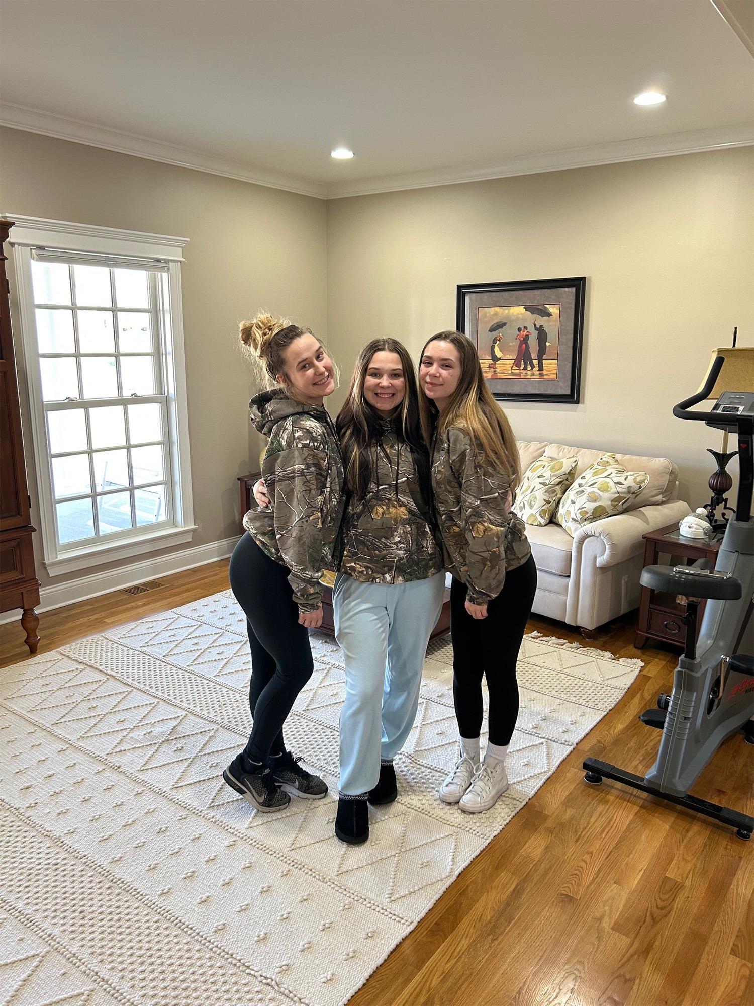 Three young women pose together in casual attire, wearing matching camouflage hoodies, smiling in a cozy living room setting.